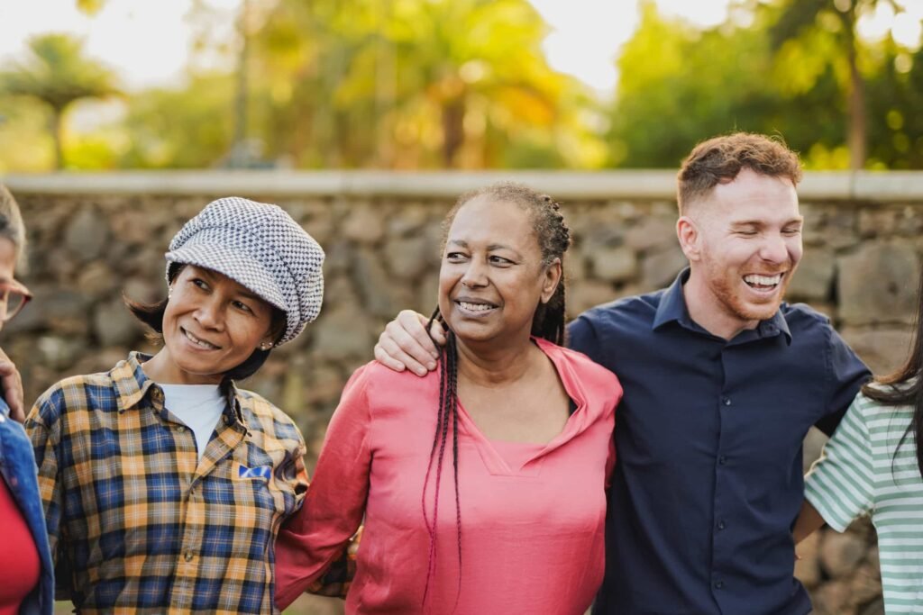 Three adults standing outdoors, smiling with arms around each other, embody the spirit of Community Sponsors. A stone wall and trees are visible in the background.