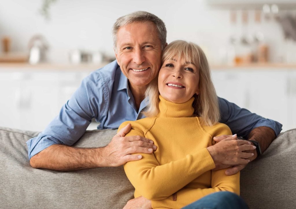 Smiling older couple sitting on a sofa at home, with the man embracing the woman from behind. Both appear happy and relaxed, enjoying peace of mind thanks to flexible financing options. Referral