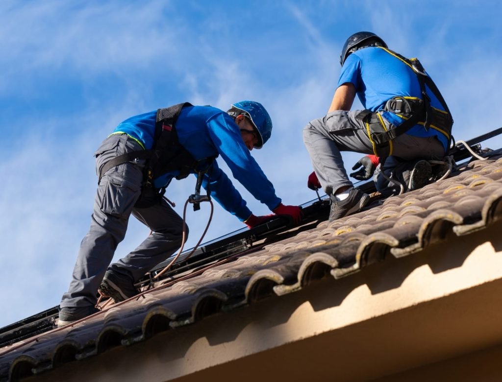 Two workers in safety gear install or repair a tiled roof on a house under a blue sky.
