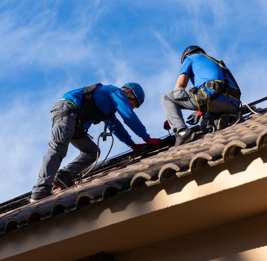 Two workers in safety gear install or repair a tiled roof on a house under a blue sky.
