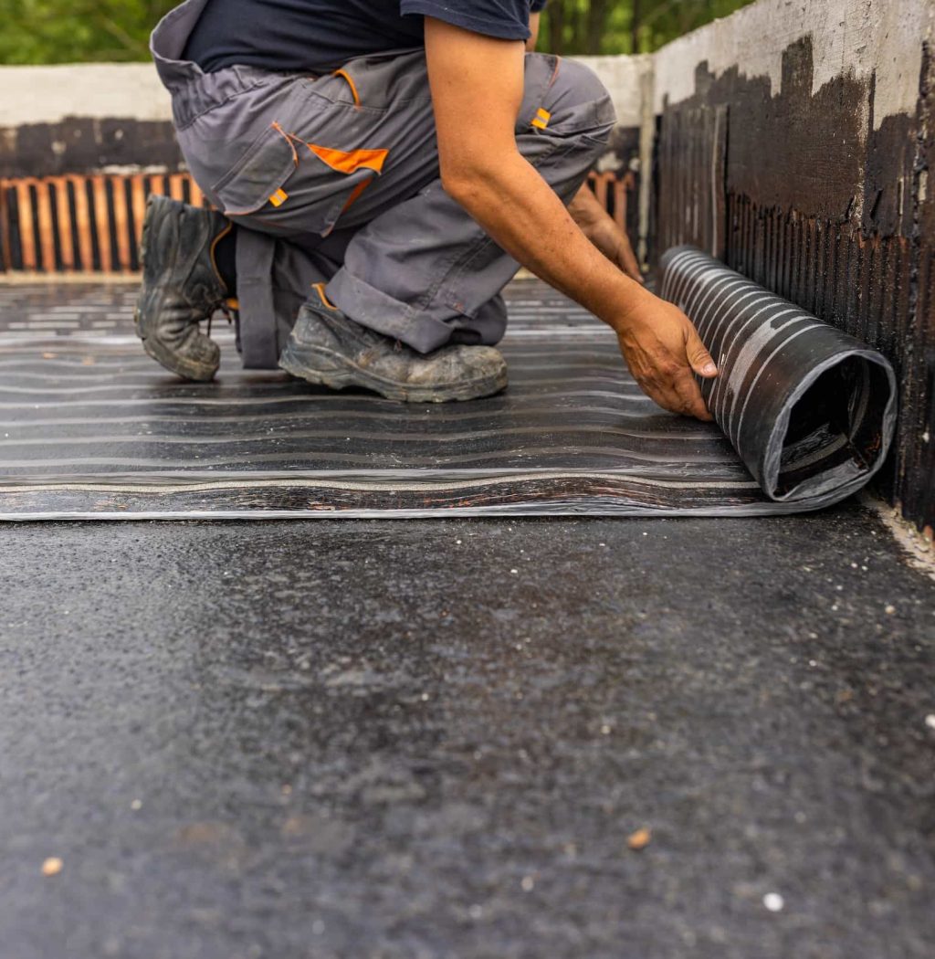 A worker kneels while unrolling a sheet of underlayment waterproofing material onto a flat concrete surface for construction or roofing.