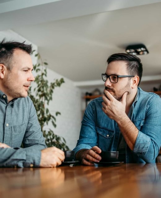 Two men sit at a wooden table indoors, engaged in conversation about the Referral Program. One man speaks while the other listens intently with his hand on his chin.