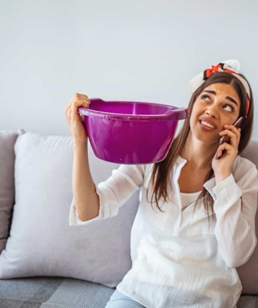 A woman sits on a couch holding a purple bucket up, looking at the ceiling, and talking on the phone, likely calling for leak detection after noticing the water issue.