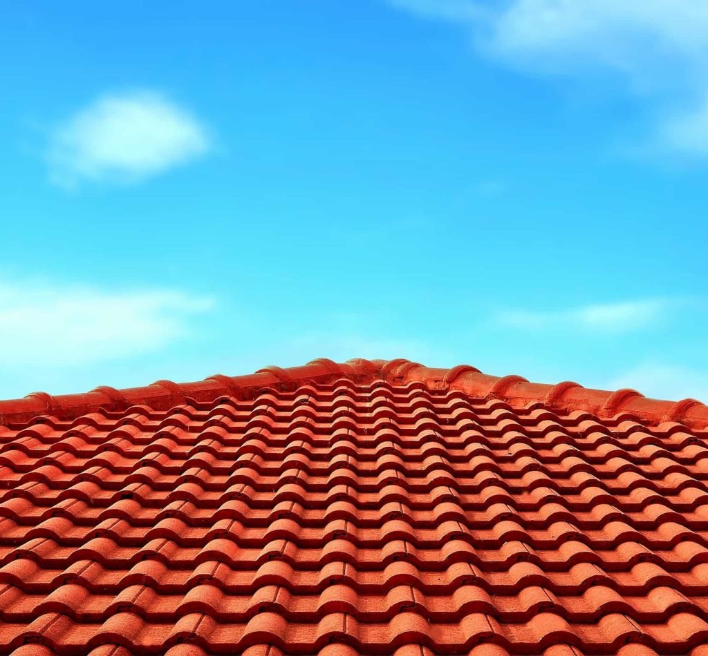 A red clay tiled roof with repeating wavy patterns is shown against a bright blue sky with a few scattered clouds.
