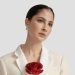 A woman in a cream blazer with a red rose pinned to the lapel, wearing silver earrings, looks slightly to the side against a plain background.